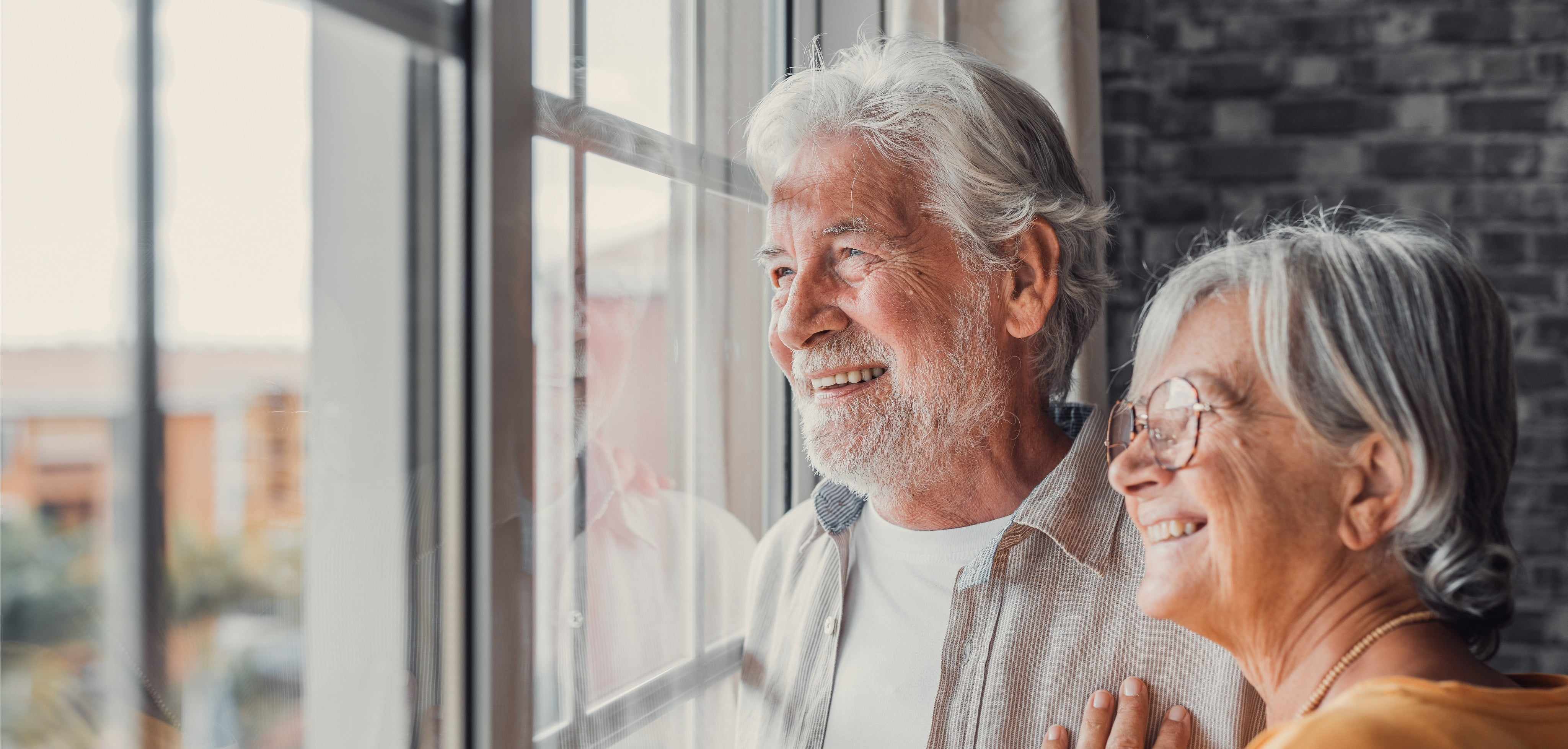 an elderly couple looking out a window smiling.