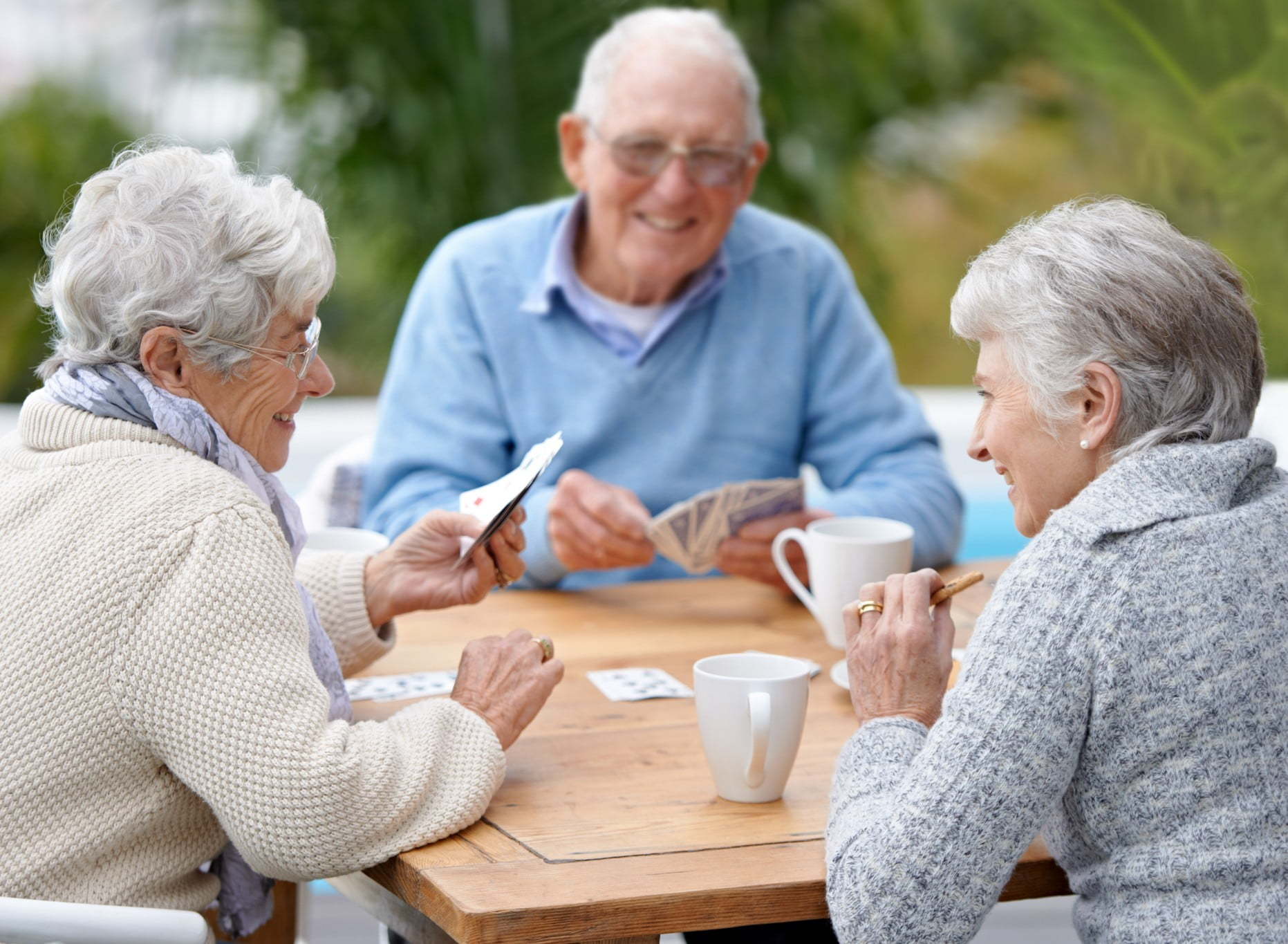 A group of elderly people laughing at a table playing cards and having coffee together.
